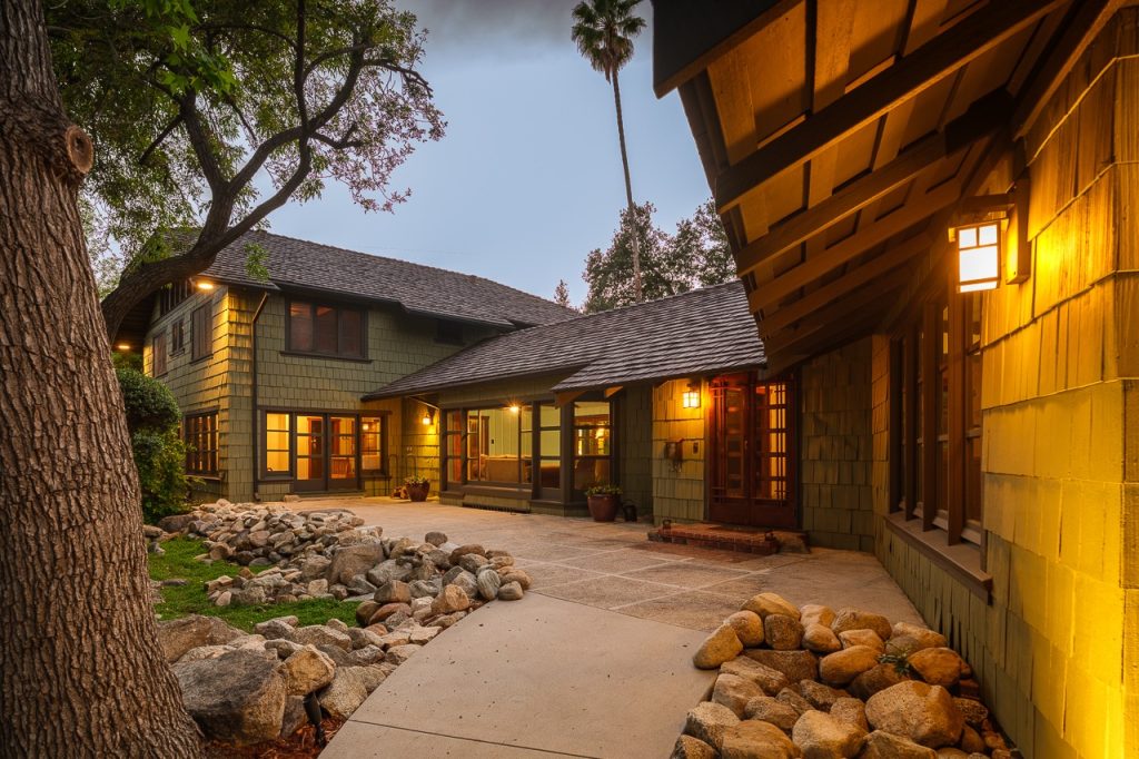 Gallery of three images showing the Edgar W. Camp House backyard: the dramatic boulder chimney with warm interior lighting, an evening view of the courtyard with extensive boulder landscaping, and the covered pergola terrace with stone paving and filtered sunlight.