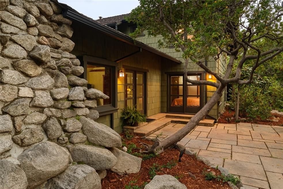 Gallery of three images showing the Edgar W. Camp House backyard: the dramatic boulder chimney with warm interior lighting, an evening view of the courtyard with extensive boulder landscaping, and the covered pergola terrace with stone paving and filtered sunlight.