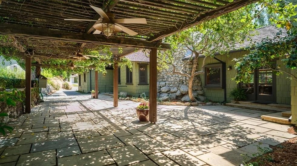 Gallery of three images showing the Edgar W. Camp House backyard: the dramatic boulder chimney with warm interior lighting, an evening view of the courtyard with extensive boulder landscaping, and the covered pergola terrace with stone paving and filtered sunlight.