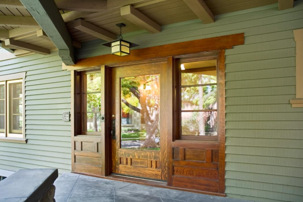 The front entrance of the Samuel P. Sanborn House showing custom wooden doors with glass panels flanked by sidelights under a covered porch with exposed beam ceiling.