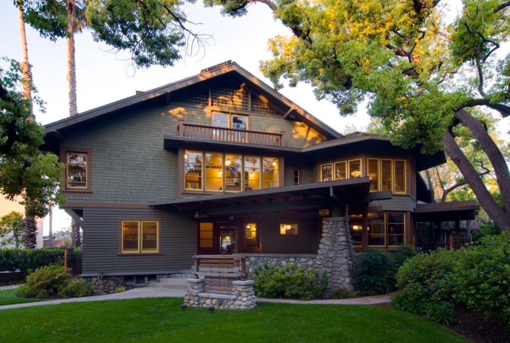 Front elevation of the Samuel P. Sanborn House at dusk showing the complete facade with stone foundation, shingled walls, multiple roof levels, and warm interior lighting visible through numerous windows.