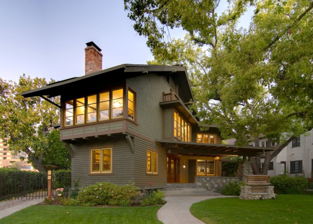Evening exterior view of the Samuel P. Sanborn House showing the two-story Craftsman residence with shingled siding, multiple gabled roofs, and extensive casement windows illuminated from within.