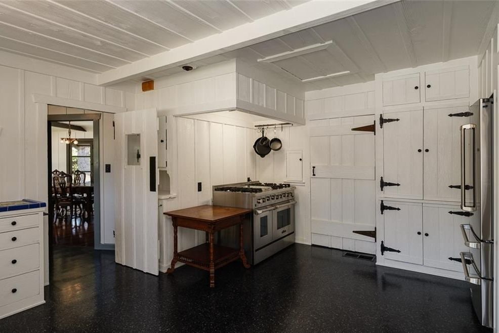 Gallery of two images showing the Edgar W. Camp House kitchen areas: a main kitchen with white board-and-batten cabinetry, exposed beam ceiling, and period hardware, and a pantry featuring dark green cabinetry with glass-front upper cabinets and exposed structural beams.