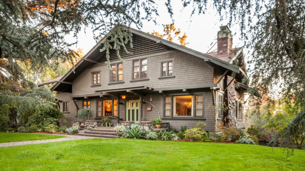 Twilight view of Lucretia Garfield house showing two-story Craftsman with wood shingles, brick chimney, and lush lawn