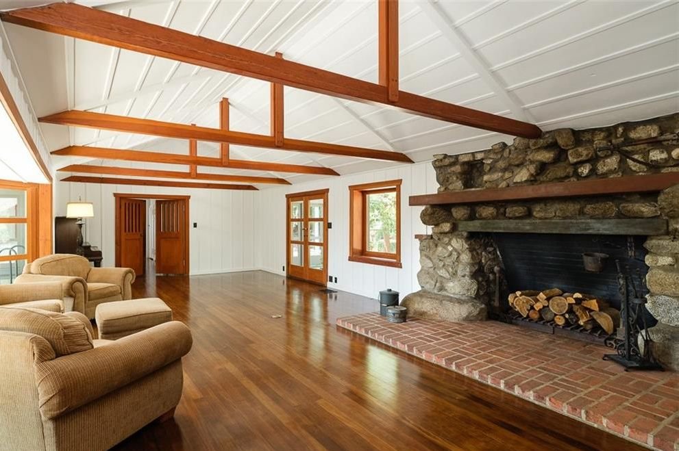 Gallery of two images showing the Edgar W. Camp House interior: a historic black and white photograph of the original living room with massive boulder fireplace and rustic furnishings, and a contemporary view of the same space showing the restored exposed beam ceiling and stone fireplace with modern furnishings.