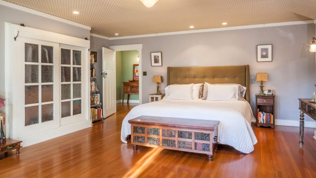 Master bedroom with tufted tan headboard, white linens, decorative storage bench, hardwood floors, and French door closets