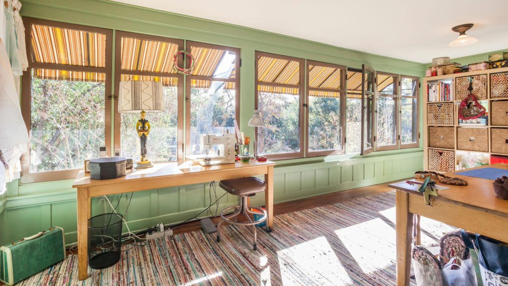 Sunlit office with green board-and-batten wainscoting, wall of windows with striped awnings, sewing machine, and craft tables