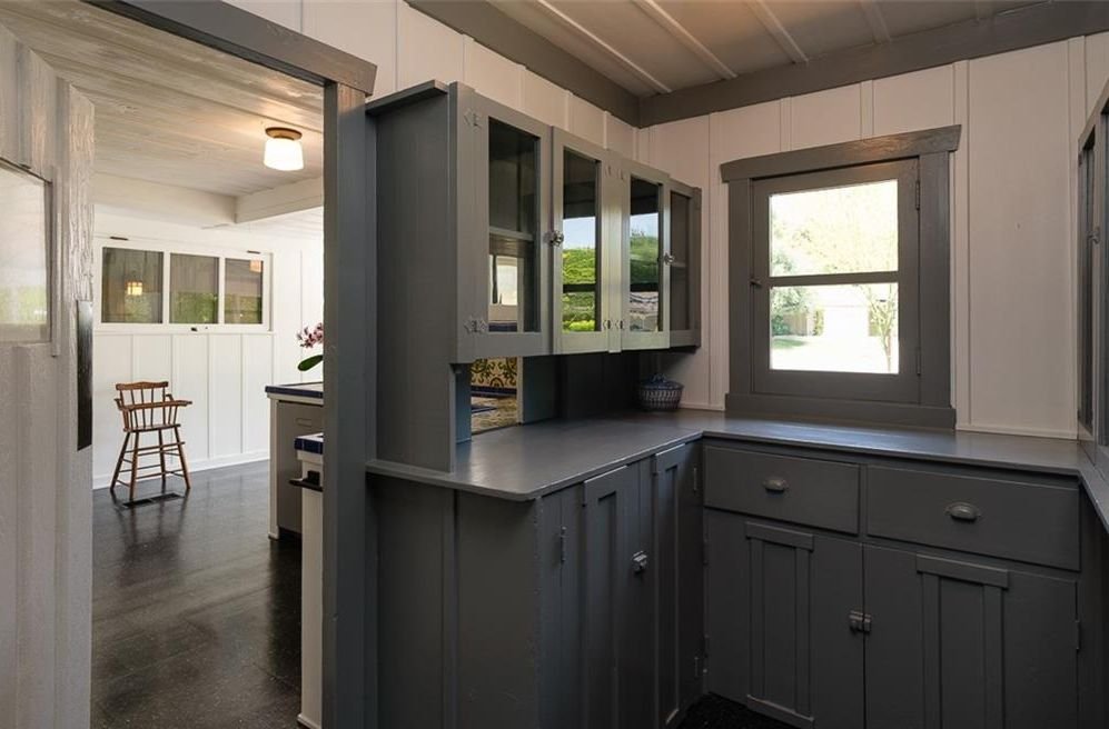 Gallery of two images showing the Edgar W. Camp House kitchen areas: a main kitchen with white board-and-batten cabinetry, exposed beam ceiling, and period hardware, and a pantry featuring dark green cabinetry with glass-front upper cabinets and exposed structural beams.