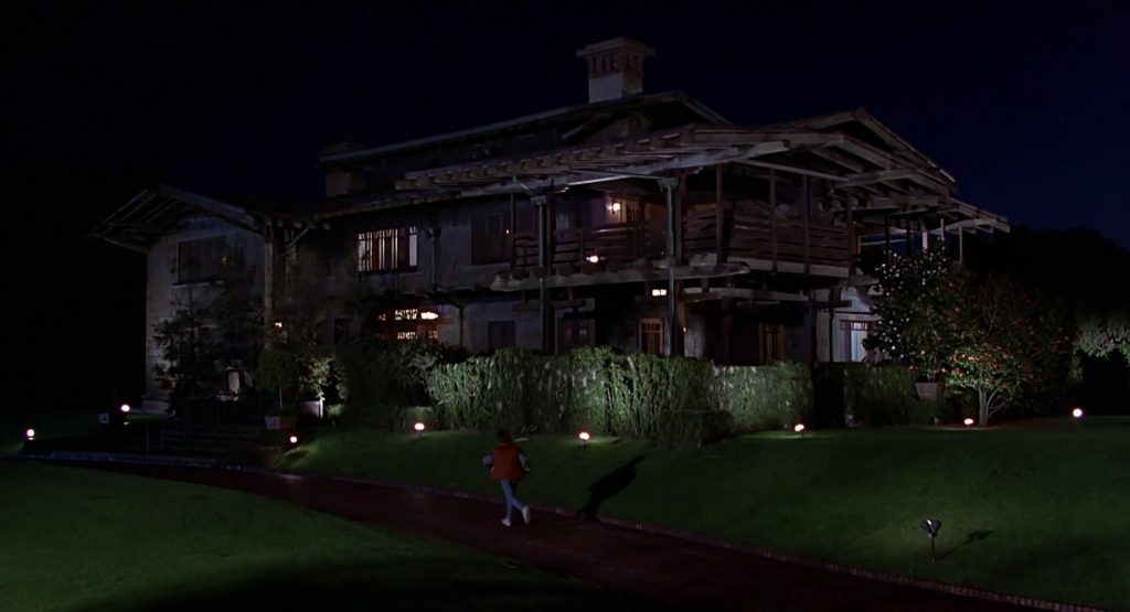 Night exterior shot from "Back to the Future" showing the illuminated Gamble House with its distinctive low-pitched rooflines, deep eaves, and sleeping porches, as Marty McFly runs across the front lawn toward the house