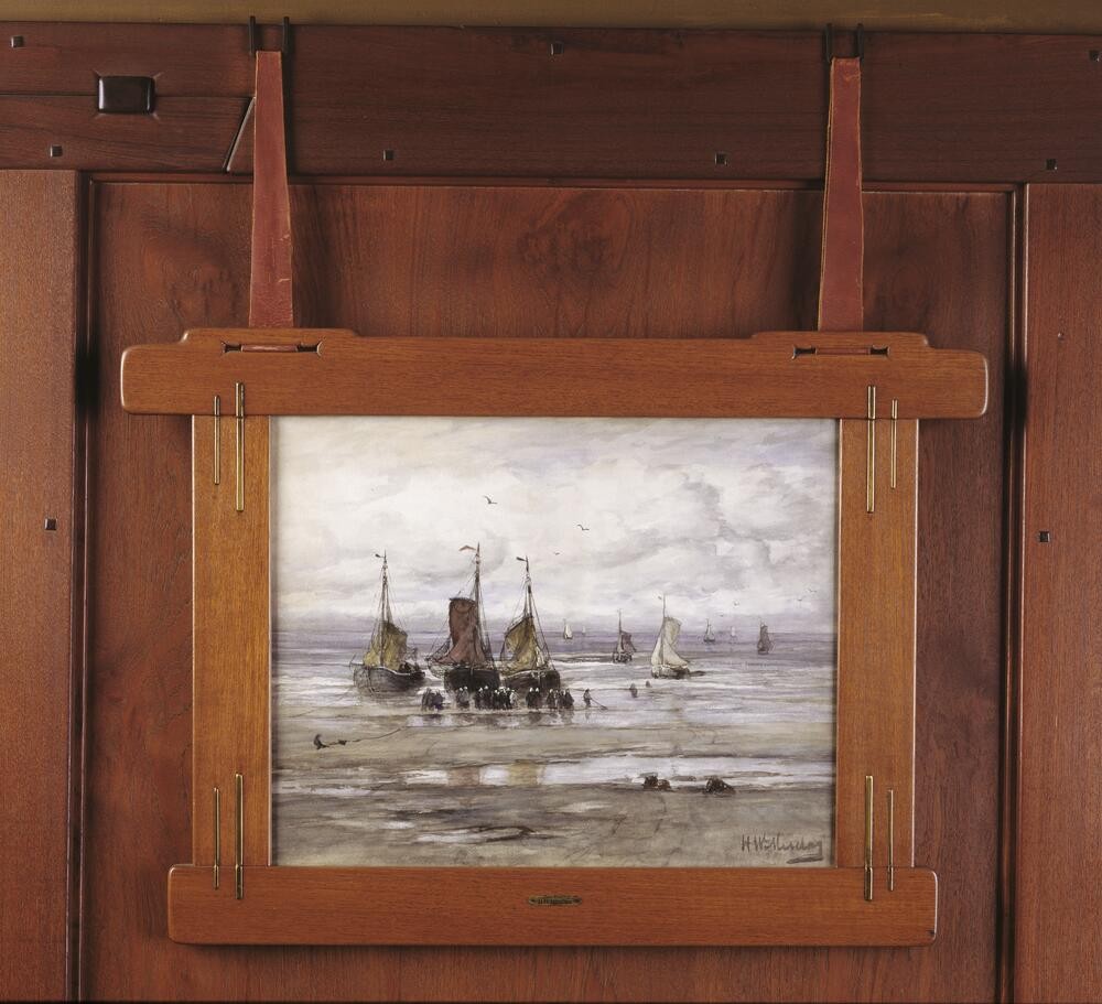Honduras mahogany picture frame with brass rod detailing holding maritime painting of fishing boats on shore. The custom Greene and Greene frame features rounded corners, visible pegging, and vertical brass rods at corners. Mounted against rich wood paneling, demonstrating integration of artwork into architectural design.
