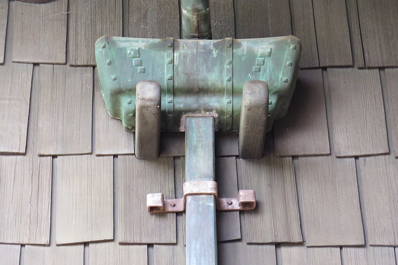 Custom copper downspout detail at the Gamble House showing the patinated green metal gutter bracket with decorative square pegs and straps securing the downspout to the weathered wood shingle siding, exemplifying Greene and Greene's attention to functional elements as ornamental features