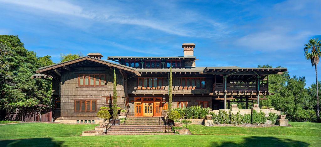 Full east facade view of the David B. Gamble House showing the asymmetrical composition with multiple gabled rooflines, second-story sleeping porches with horizontal slat railings, projecting eaves with exposed rafter tails, weathered wood shingle siding, brick terrace steps, and clinker brick retaining walls, set against a clear blue sky and manicured lawn