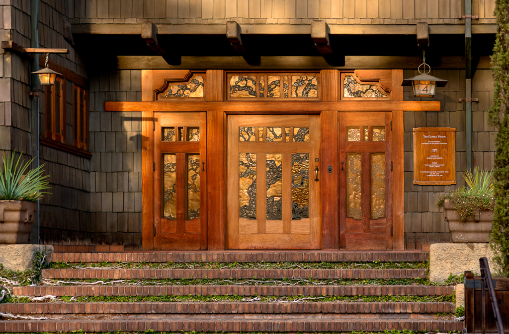 Twilight view of the Gamble House main entrance showing the triple-door configuration with glowing art glass panels depicting stylized tree branches, flanked by Arts and Crafts lanterns under deep eaves supported by exposed timber brackets, with brick steps leading to the teak entry doors