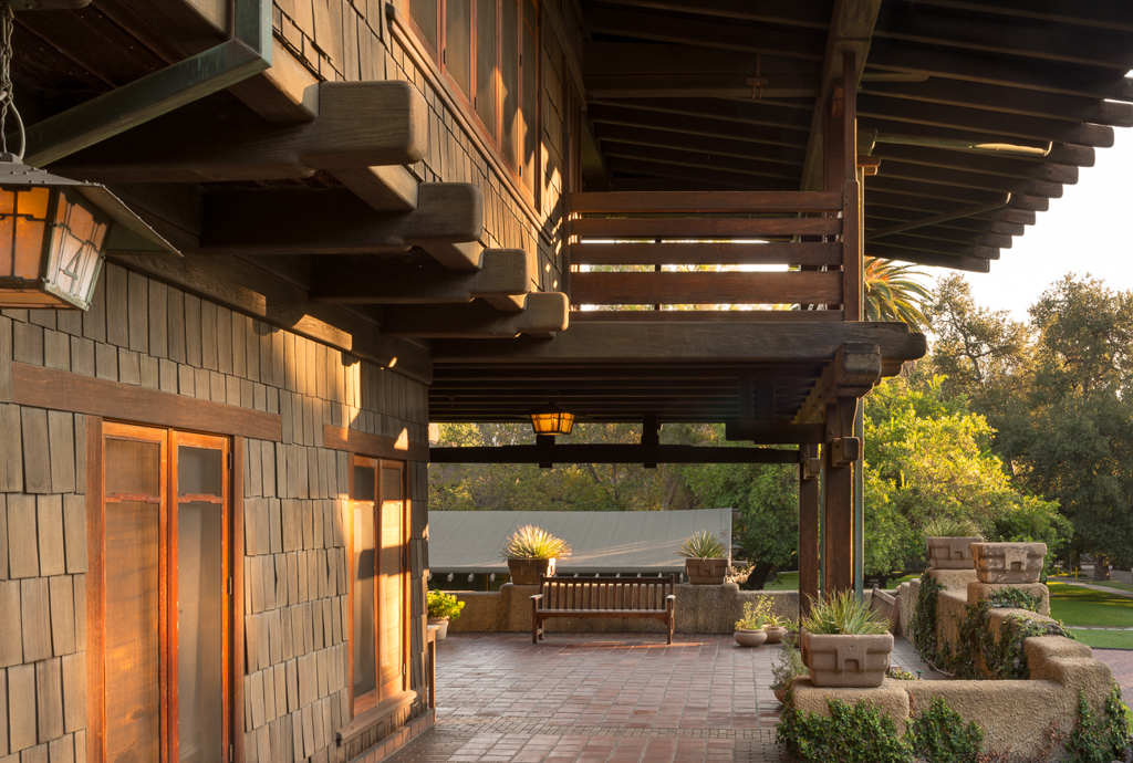 Front terrace of the Gamble House at sunset showing the brick-paved outdoor living space with a built-in teak bench between clinker brick planters, covered by a massive timber sleeping porch with exposed beam construction and Arts and Crafts lanterns, overlooking the landscaped grounds