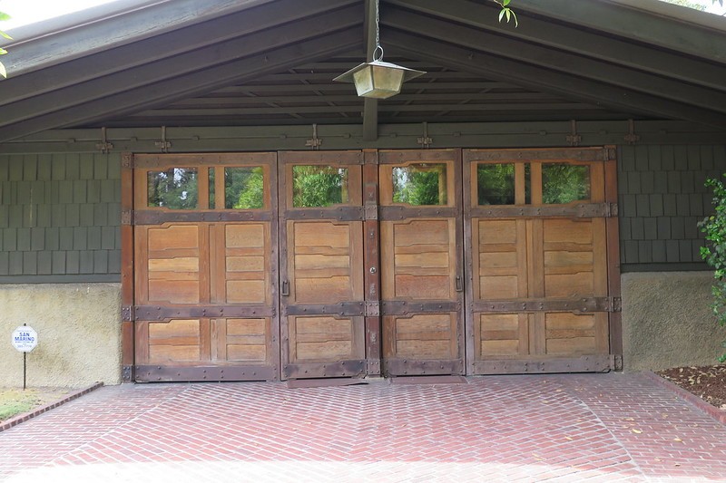 The Gamble House garage with its full bank of wooden doors closed, frame-and-panel construction below and glazed lights above, beneath a broad shingled gable roof with a lantern hanging at the peak
