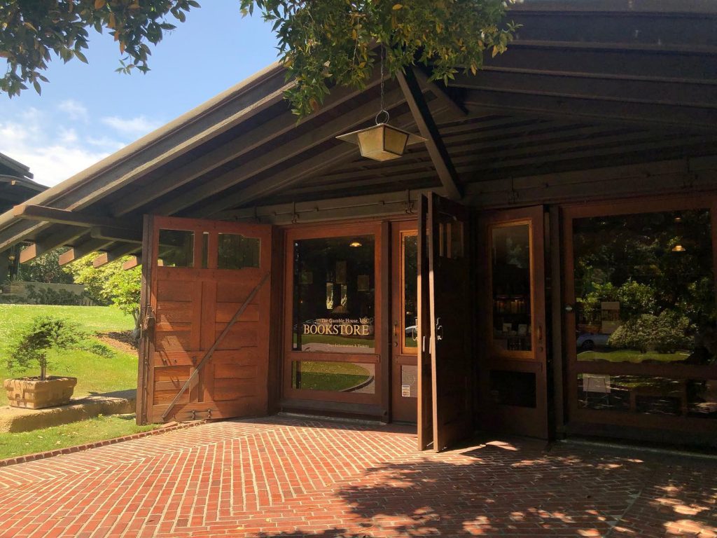 Gamble House garage with bay doors open revealing the current use as the Gamble House Bookstore, showing the teak wood doors with glass panels, exposed timber eave construction with a hanging lantern, and herringbone brick paving leading to the entrance, surrounded by mature trees and landscaping