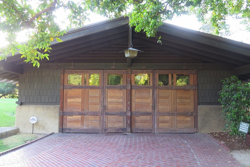 Front view of the Gamble House garage showing six custom teak wood garage doors with horizontal panel designs and small square windows at the top, set beneath a low-pitched gable roof with exposed rafter tails, wood shingle siding above a stucco base, and a herringbone brick driveway, with an Arts and Crafts lantern hanging from the eave