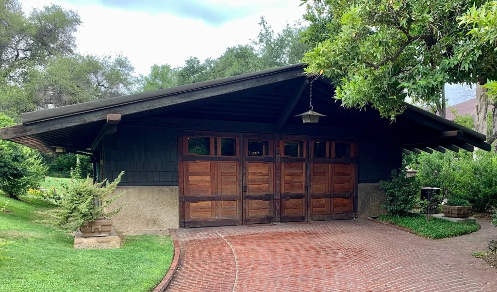 Front facade of the Gamble House garage showing the six-bay configuration with custom teak wood doors featuring horizontal panels and upper glass lights, weathered wood shingle siding, stucco base walls, deep overhanging eaves with exposed structural elements, curved herringbone brick driveway, and an Arts and Crafts lantern suspended from the roof peak