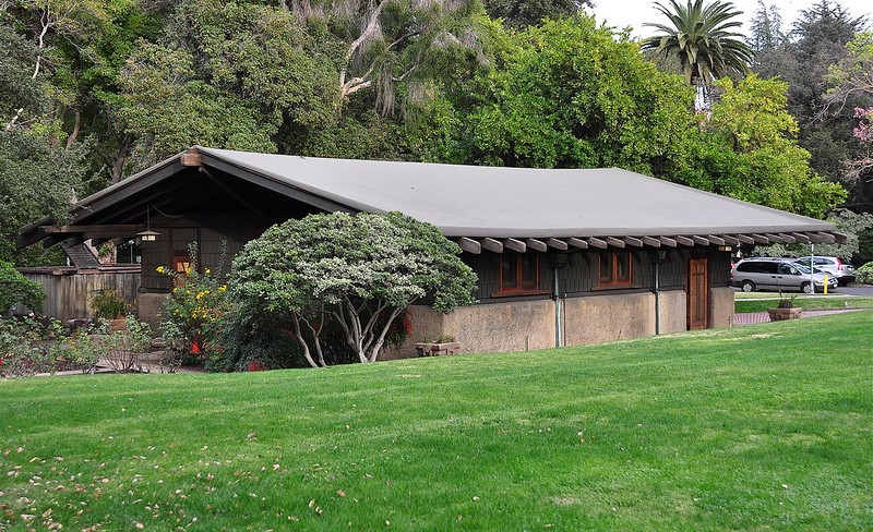 Rear elevation of the Gamble House garage showing the long low-pitched roofline with exposed rafter tails and decorative brackets, stucco walls with wood-trimmed windows, wood shingle siding at the gable ends, and the structure nestled into the sloping site with mature landscaping including flowering shrubs and specimen trees, with the street visible in the background