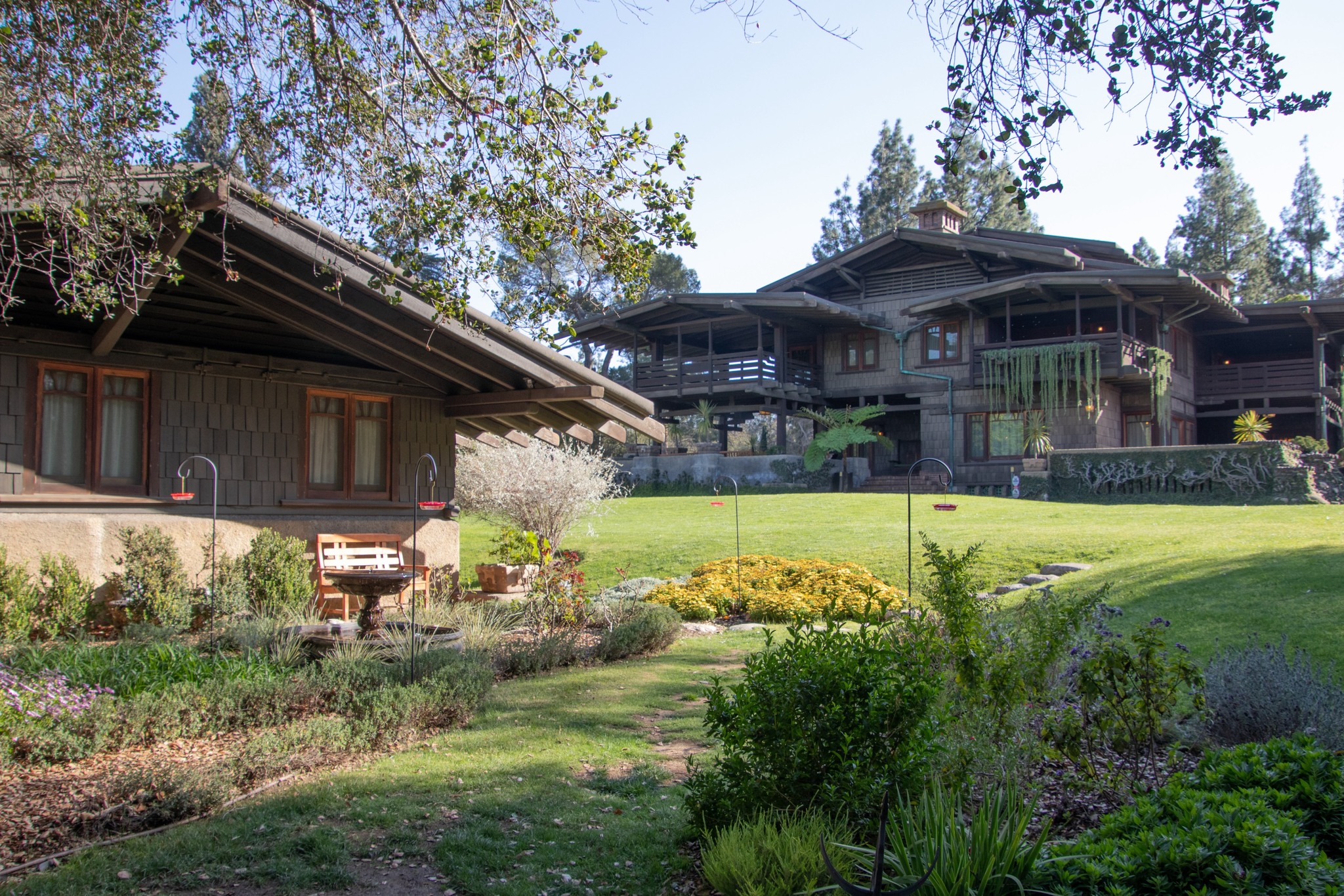 The Gamble House garage seen across the rear garden with the main house rising behind it, both buildings sharing the same shingle cladding and broad overhanging rooflines