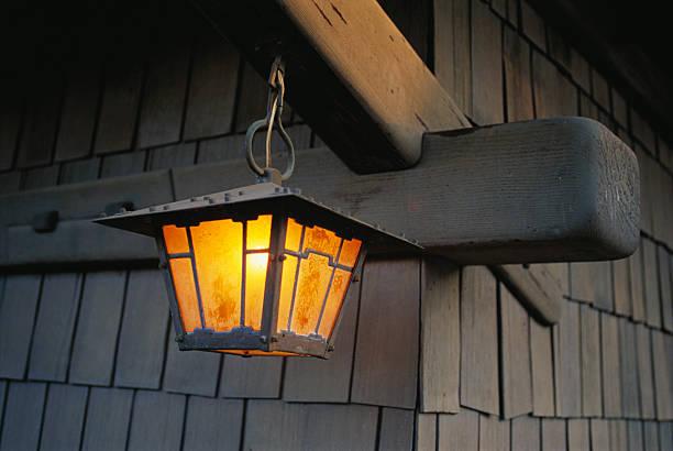 Close-up detail of an Arts and Crafts lantern at the Gamble House showing the custom-designed fixture with a dark patinated metal frame in a tapered square form, amber stained glass panels with geometric leading, suspended from a curved wrought iron bracket attached to a massive curved timber eave support, with weathered wood shingle siding visible in the background, illuminated at twilight