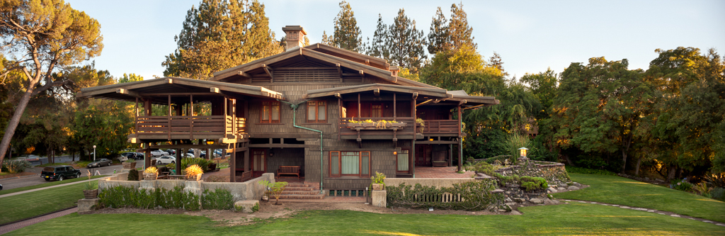 Panoramic aerial view of the Gamble House north facade at golden hour showing the full symmetrical composition with sleeping porches flanking the central entrance, the complex layering of low-pitched gabled roofs with exposed rafter tails, brick chimney, timber pergola structures, terraced clinker brick and boulder retaining walls with plantings, the curved driveway visible at left, mature oak and eucalyptus trees framing the property, and the warm glow of sunset light illuminating the weathered wood shingle siding and teak woodwork