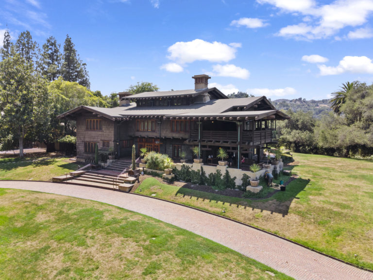 Aerial drone photograph of the Gamble House from the northeast showing the complete composition of the main house with its asymmetrical arrangement of multiple gabled roofs, central brick chimney, second-story sleeping porches on both wings, exposed rafter tails, the front terrace with clinker brick retaining walls and steps, curved concrete walkways leading to the entrance, manicured lawns, mature specimen trees including palms and eucalyptus, and the San Gabriel Mountains visible in the background under a partly cloudy sky