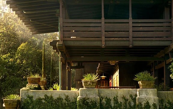 North terrace of the Gamble House photographed by Alexander Vertikoff showing the front entrance viewed from below, with the second-story sleeping porch featuring horizontal slat railings, massive timber posts and beams supporting the deep overhanging eaves, clinker brick retaining walls with moss and patina, concrete planters with agave and ornamental grasses, and the teak entrance doors visible in the shadowed recessed entry, with dappled sunlight filtering through mature trees