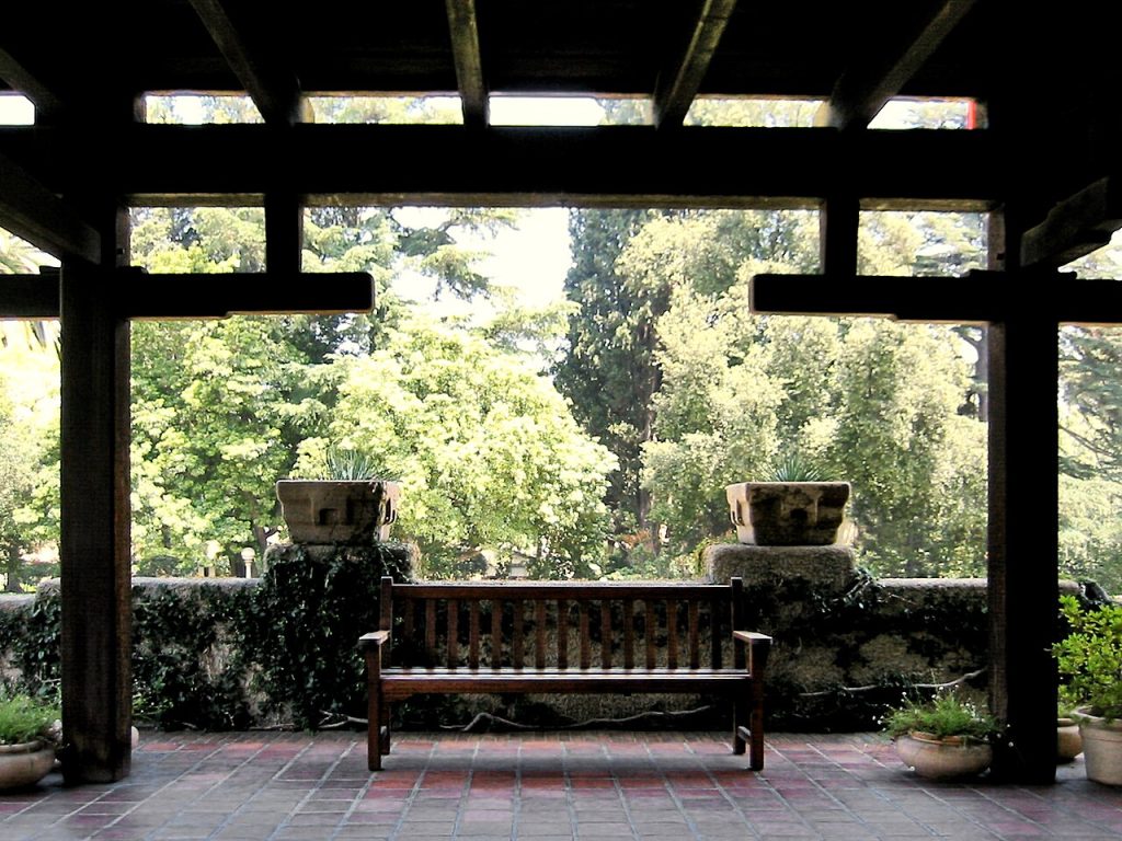 View from beneath the north terrace sleeping porch looking out toward the garden, showing the silhouette of massive timber beams and posts framing the view, a custom Greene and Greene teak bench with horizontal slat back centered on the brick-paved terrace between two concrete planters with weathered patina, and the lush garden landscape with mature trees visible beyond, creating a contemplative outdoor room that connects the house to its natural surroundings