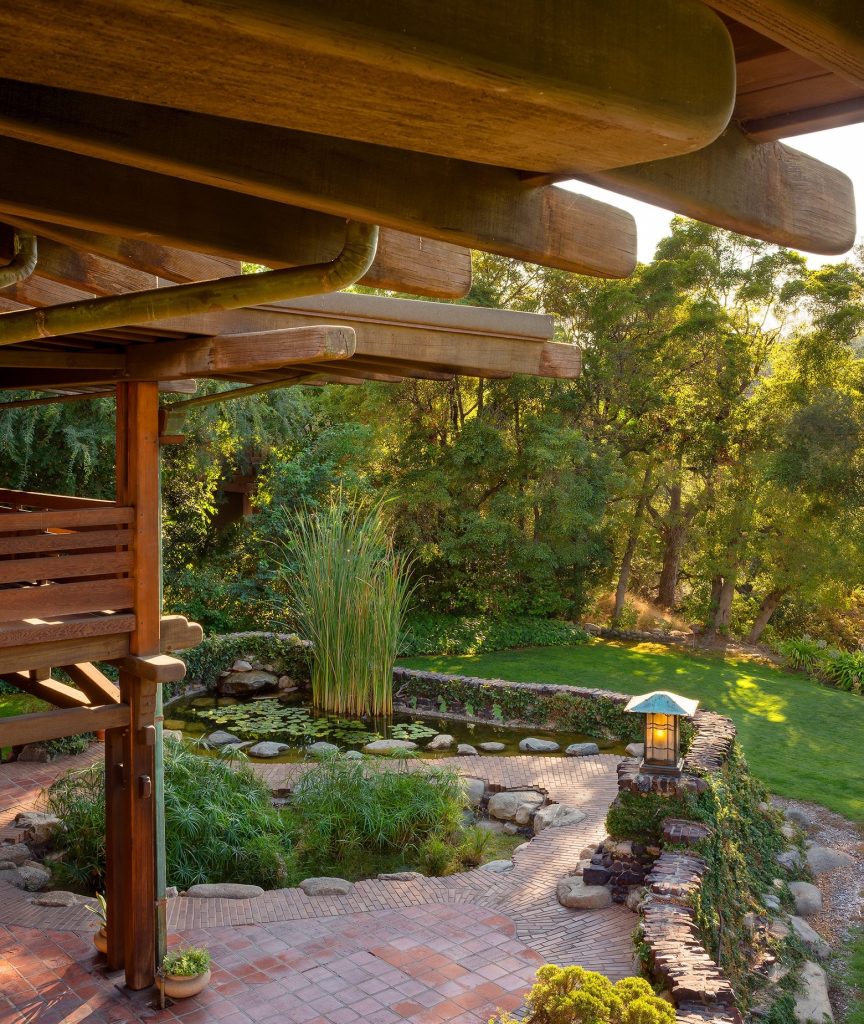 Aerial view from the Gamble's sleeping porch photographed by Alexander Vertikoff showing the rear terrace's naturalistic water garden with lily pond featuring water lilies, ornamental grasses, large boulders, and aquatic plants, surrounded by herringbone brick paving, clinker brick and stone retaining walls with moss and patina, an Arts and Crafts lantern on a brick pedestal, and the massive timber pergola structure overhead with rounded rafter tails and exposed beam construction, with the manicured lawn and mature trees visible beyond in golden afternoon light