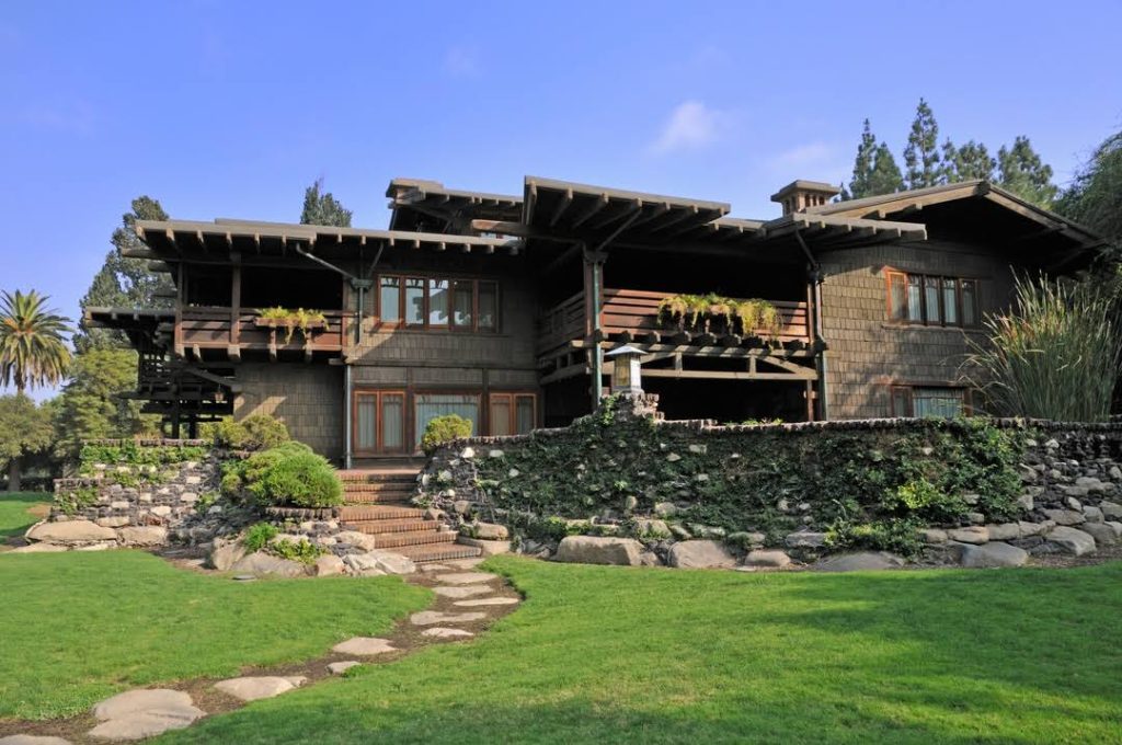 Rear view of the Gamble House from the backyard showing the south facade with multiple levels of sleeping porches featuring horizontal slat railings, central brick chimney with decorative cupola, complex arrangement of low-pitched gabled roofs with exposed rafter tails and decorative brackets, weathered wood shingle siding, terraced clinker brick and boulder retaining walls heavily planted with ivy and ground cover, brick steps ascending through the landscaping, curved flagstone pathway through the manicured lawn, and mature palm and eucalyptus trees framing the composition under a bright blue sky
