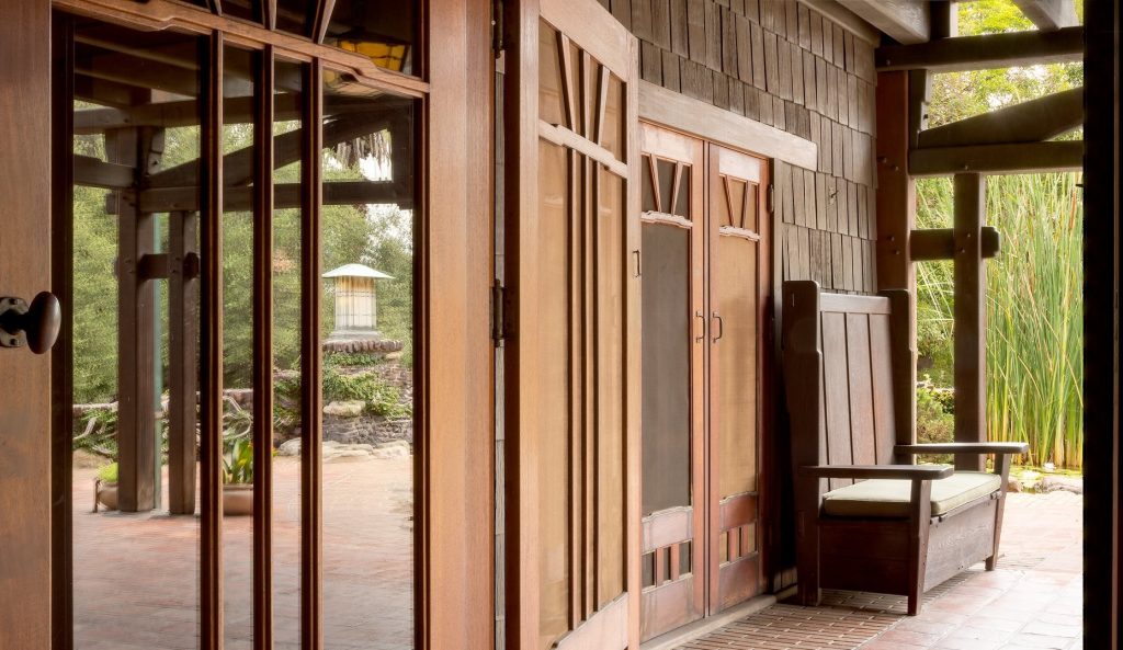View from just inside the rear doors leading to the rear terrace photographed by Alexander Vertikoff showing custom Greene and Greene designed teak storage cabinets with geometric panel designs and V-shaped muntins in the upper glass doors, a teak bench with horizontal slats at right, massive timber posts and beams framing the space, weathered wood shingle siding, brick paving, and views through the open colonnade to the naturalistic garden with lily pond, Arts and Crafts lantern on brick pedestal, and ornamental grasses beyond