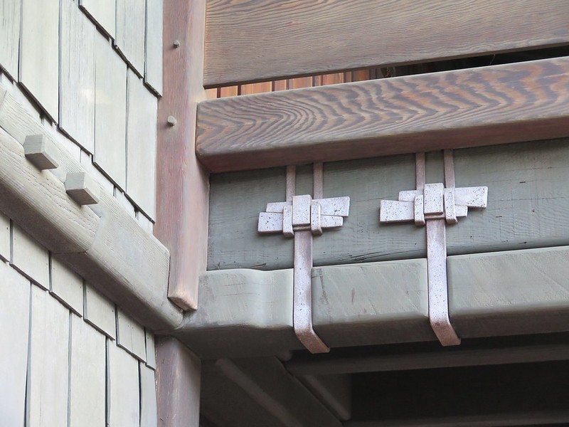 Close-up architectural detail of Greene and Greene's signature timber joinery at the Gamble House showing wooden straps with proud square pegs securing a massive rounded beam to vertical posts, with weathered wood shingle siding and painted horizontal siding visible, demonstrating the exposed structural system and handcrafted joinery that characterizes the ultimate bungalow aesthetic where functional elements become decorative features