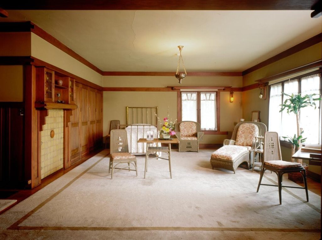 Aunt Julia Huggins' bedroom at the Gamble House furnished with soft green painted wicker furniture including high-backed chairs with vertical slat designs, a chaise lounge, side chairs, and occasional table, arranged on a cream-colored area rug with geometric border pattern, with a built-in fireplace featuring cream-colored tile surround and Honduras mahogany mantel and cabinetry at left, windows with V-shaped muntins and sheer curtains providing natural light, cream-colored walls with mahogany trim, exposed beam ceiling, hanging pendant light fixture, and fresh flowers on the table, creating a light and airy feminine space distinct from the darker wood tones found elsewhere in the house