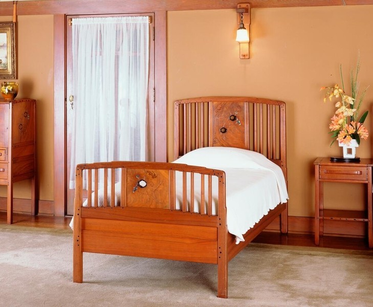 Master bedroom featuring Greene and Greene-designed bed in Honduras mahogany with vertical slat headboard and footboard. Central decorative panel displays abalone and mother-of-pearl inlay. Room includes matching nightstand, wall sconce, and period furnishings in warm Arts and Crafts interior setting.