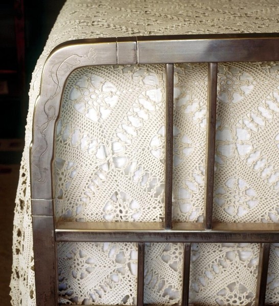 Detail of nickel-plated brass bedframe showing curved corner joint with engraved floral decoration. Vertical slats frame vintage crocheted lace coverlet. The metalwork demonstrates Greene and Greene's collaboration with skilled artisans and attention to decorative details in utilitarian furniture components.