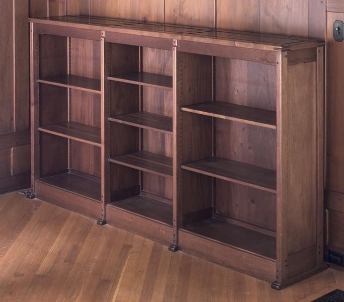 Expandable bookcase system in Honduras mahogany with three sliding sections on metal casters. Features adjustable shelving, visible joinery details, and modular design allowing flexible configuration. Photographed in fully extended position against wood-paneled wall, demonstrating innovative functional furniture design.