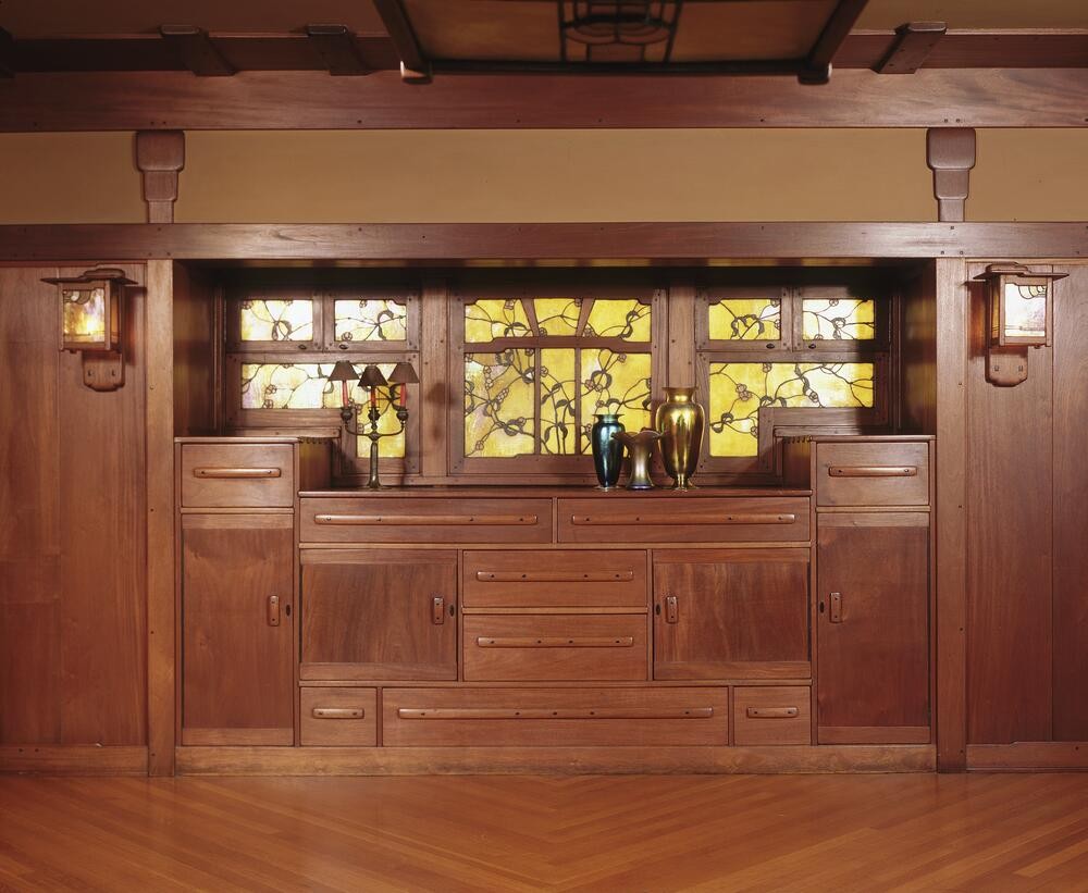 Custom-designed built-in sideboard in the Gamble House dining room crafted by Peter Hall Manufacturing Company, featuring Honduras mahogany construction with multiple drawers and cabinet doors with copper hardware, flanked by Arts and Crafts wall sconces, with backlit Tiffany Studios stained glass panels depicting stylized oak branches with golden-yellow glass set within the upper cabinet doors, displayed with period pottery including iridescent green and amber vases, beneath exposed timber beam construction with cloud-lift brackets and a decorative lantern, demonstrating Greene and Greene's integration of architecture, furniture, and decorative arts