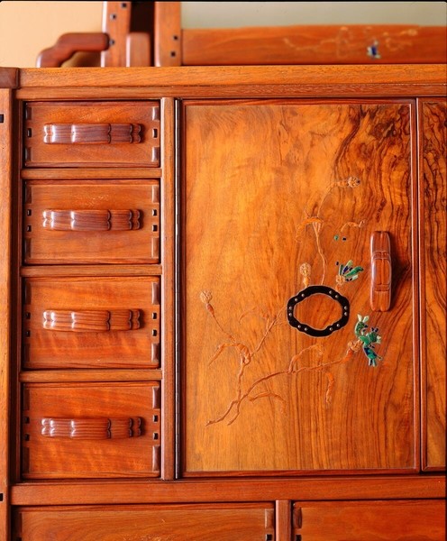 Overhead detail view of a Greene and Greene designed chiffonier at the Gamble House showing the rich figured black walnut wood grain on the cabinet door, with decorative silver and abalone shell inlay depicting stylized oak leaves and acorns in an organic curvilinear pattern, flanked by drawers with custom copper hardware featuring cloud-lift pulls, and proud square ebony pegs securing the construction, exemplifying the brothers' integration of natural motifs and precious materials