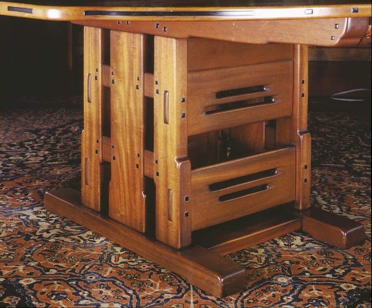 Detail view of the Greene and Greene designed dining table pedestal base at the Gamble House, photographed from below showing the complex structural system with massive Honduras mahogany legs featuring vertical panels with horizontal slat cutouts, square ebony pegs throughout, copper and leather strap hardware, curved stretchers connecting the legs, and the table resting on an Arts and Crafts carpet with floral patterns, demonstrating the sculptural quality and exposed joinery that characterizes the brothers' furniture design