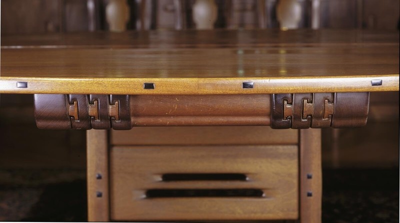 Close-up detail of the Greene and Greene designed dining table at the Gamble House showing the underside construction with Honduras mahogany top, with square ebony pegs and splines, custom copper and leather strap hardware securing the apron to the legs, and a drawer with cloud-lift copper pulls, demonstrating the brothers' meticulous attention to joinery details and decorative hardware even on hidden structural elements