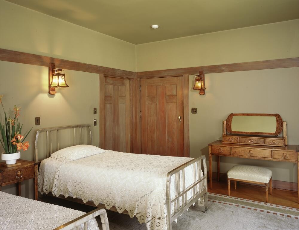 Guest bedroom at the Gamble House showing twin metal beds with vertical slat headboards and footboards painted in soft green, dressed with white crocheted bedspreads, flanked by a Honduras mahogany nightstand with copper hardware, a dressing table with tilting mirror and upholstered bench at right, custom Honduras mahogany pocket doors with geometric panel design, Arts and Crafts wall sconces with amber glass shades, cream-colored walls with mahogany trim, exposed beam ceiling painted soft green, and a patterned area rug, creating a serene and cohesive sleeping space