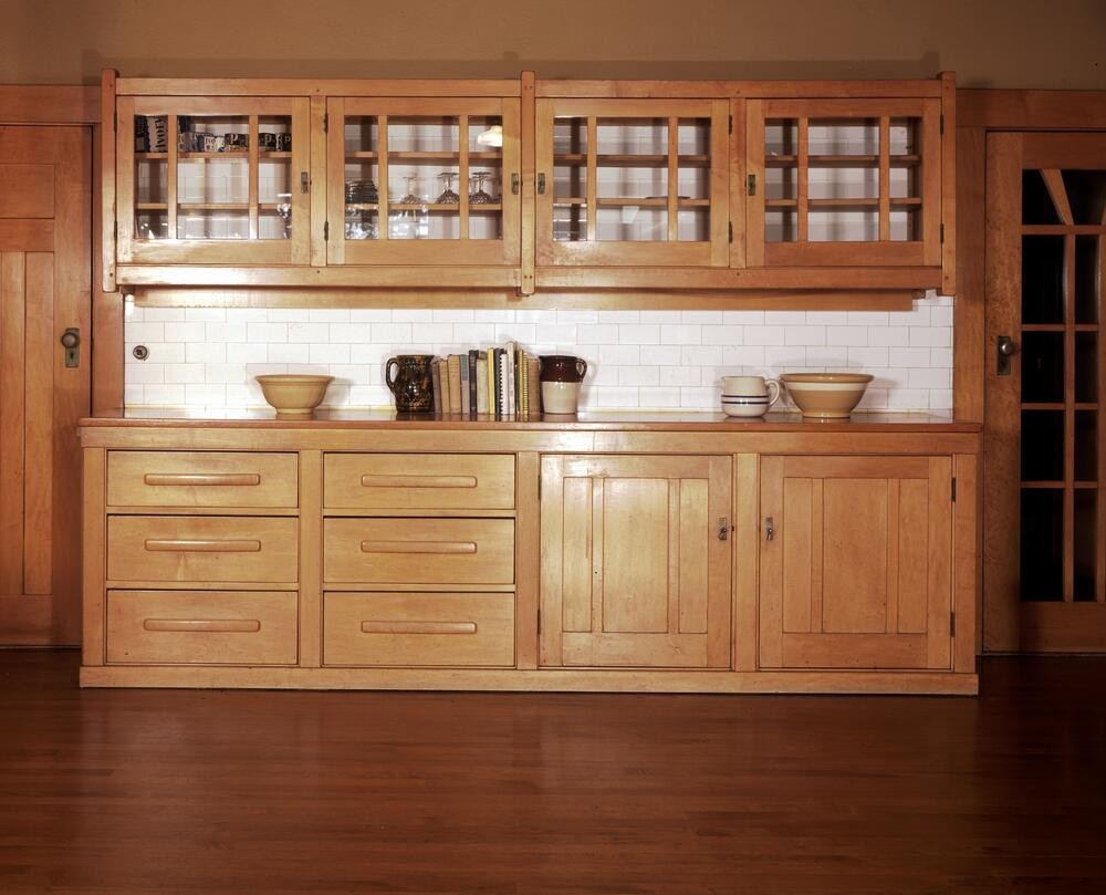 Built-in kitchen cabinetry system in maple with sugar pine countertops, featuring glass-front upper cabinets displaying dishware, multiple drawers with horizontal pulls, and lower cabinet doors. White subway tile backsplash contrasts with warm wood tones, demonstrating functional Arts and Crafts kitchen design integrated with surrounding Honduras mahogany woodwork.
