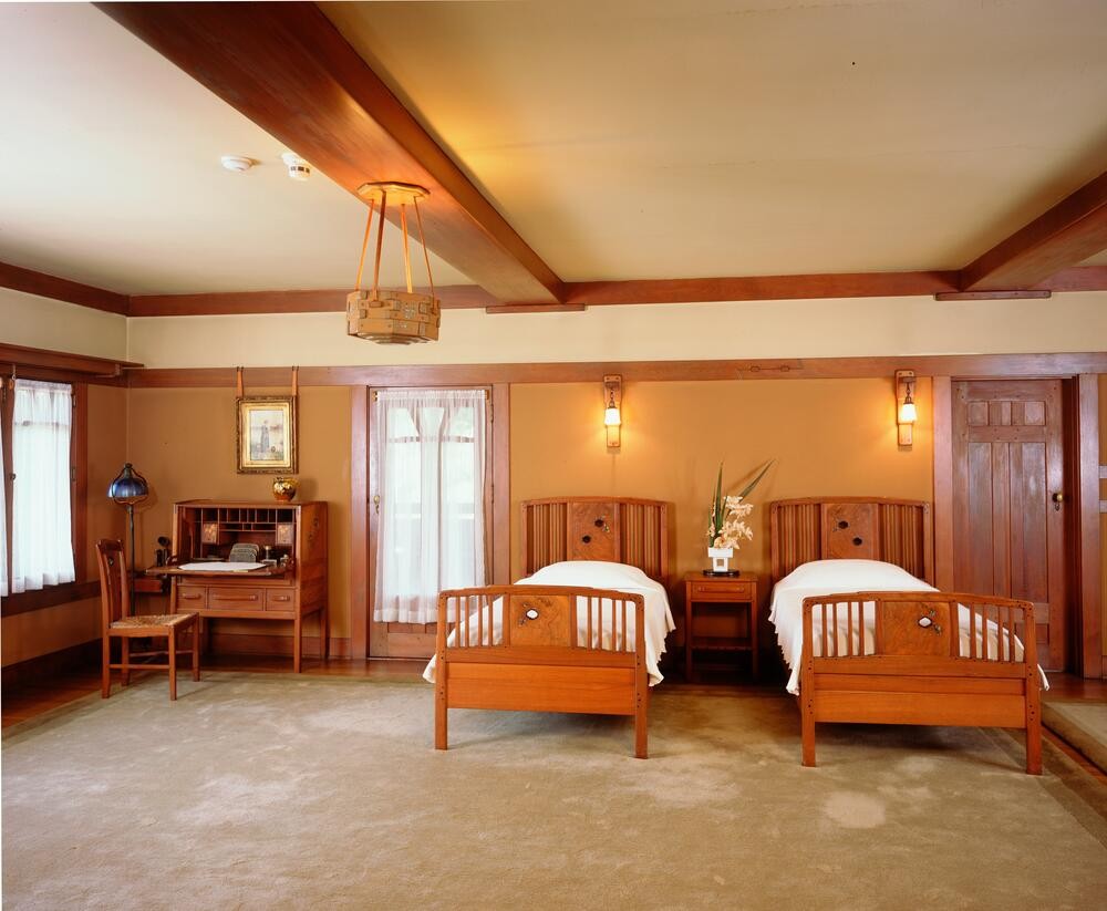 Master bedroom at the Gamble House showing twin Honduras mahogany beds with vertical slat headboards and footboards, dressed with white coverlets, separated by matching nightstands, with a dressing table and chair at left, door to the sleeping porch at center with sheer curtains, three Arts and Crafts wall sconces with amber glass shades providing ambient lighting, custom Honduras mahogany pocket door at right, exposed beam ceiling with cream-colored plaster between mahogany beams, warm peach-toned walls with mahogany trim and wainscoting, a custom pendant light fixture with geometric design suspended from the ceiling, and cream-colored carpet, creating a warm and intimate sleeping space with the characteristic integration of architecture and furnishings
