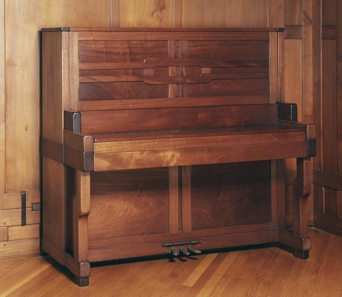 Custom upright piano in the living room of the Gamble House with case redesigned by Greene and Greene, featuring Honduras mahogany and bird's-eye maple construction with geometric panel designs on the front incorporating contrasting wood tones, exposed structural posts with square ebony pegs, black metal hardware and hinges, positioned against Honduras mahogany wall paneling with V-shaped muntins, and hardwood flooring with geometric border pattern, demonstrating the brothers' comprehensive approach to integrating even musical instruments into their total design aesthetic