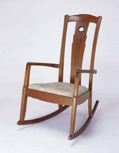 Greene and Greene designed rocking chair from the Gamble House crafted by Peter Hall Manufacturing Company in Honduras mahogany and black walnut, featuring a tall back with cloud-lift cutout handle at the crest rail, vertical splats with three linear grooves and a circular abalone shell inlay at the top, open arms with curved supports, upholstered seat in cream fabric with stylized floral pattern in soft colors, curved rockers, square ebony pegs throughout, and the characteristic rounded edges that exemplify the ultimate bungalow furniture aesthetic