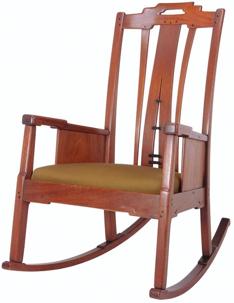Greene and Greene designed rocking chair from the Gamble House crafted by Peter Hall Manufacturing Company in Honduras mahogany with ebony pegs, featuring a tall back with cloud-lift cutout handle, vertical and horizontal slats creating a geometric pattern with square ebony pegs as decorative accents, solid side panels under the arms, upholstered seat in golden-yellow leather, curved rockers, and the exposed joinery and structural expressiveness characteristic of the brothers' furniture design