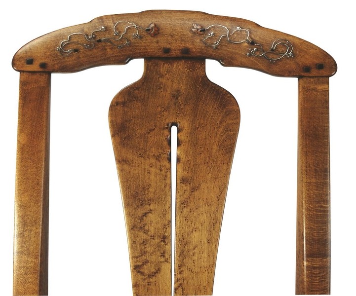 Close-up detail of the crest rail of a Greene and Greene rocking chair from the Gamble House showing the cloud-lift cutout handle decorated with silver and abalone shell inlay depicting stylized curving vine tendrils with small flowers, square ebony pegs flanking the decorative inlay, figured bird's-eye maple central splat showing the distinctive grain pattern, and the characteristic rounded edges and meticulous craftsmanship that demonstrate the brothers' integration of structural elements with precious decorative materials