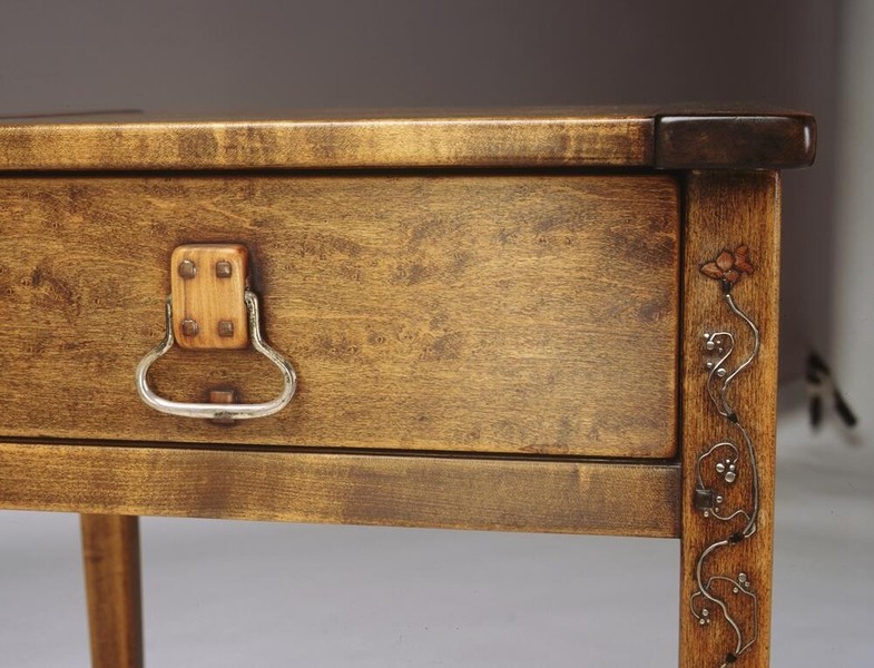 Nightstand detail in Honduras mahogany showing single drawer with nickel-plated brass pull and decorative maple inlay. Corner post features carved organic vine motif with flowing plant forms. Demonstrates Greene and Greene's integration of functional hardware with artistic embellishment and natural wood grain.