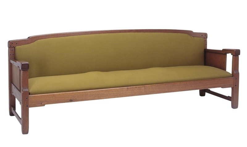 Greene and Greene designed sofa from the Gamble House crafted by Peter Hall Manufacturing Company in Honduras mahogany, featuring a long horizontal form with cloud-lift cutout handles at both ends of the crest rail, solid side panels with square ebony pegs, exposed structural posts and stretchers, and upholstered back and seat cushions in golden-yellow fabric, demonstrating the brothers' approach to seating furniture with visible structural elements and the characteristic rounded edges found throughout their ultimate bungalow designs